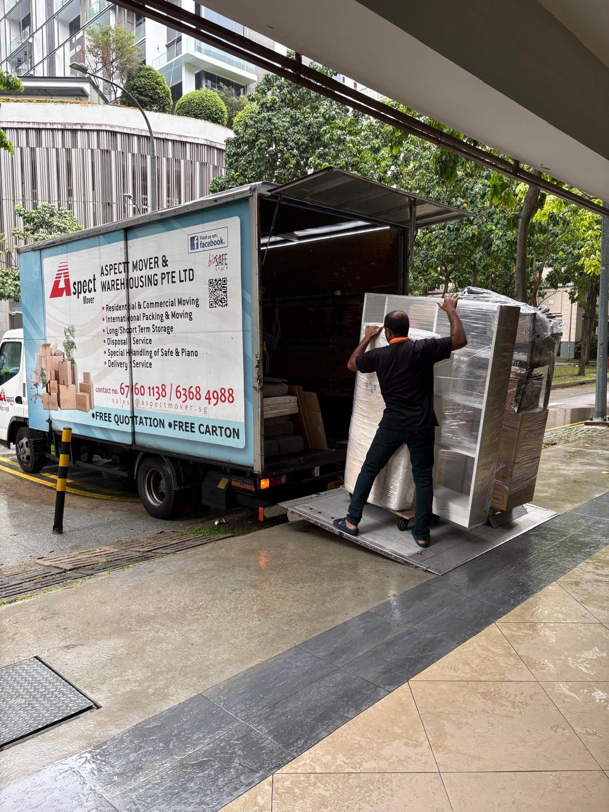 An Aspect Mover crew member seen from behind wheeling a wrapped cabinet up the tail lift of a company lorry